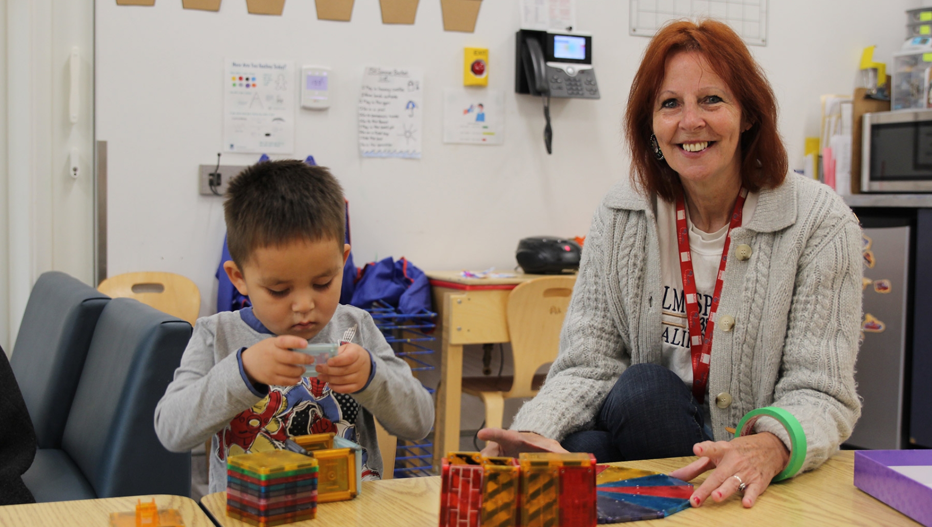 Smiling early childhood educator with shoulder-length red hair sitting next to a young boy playing with magnetic plastic blocks.